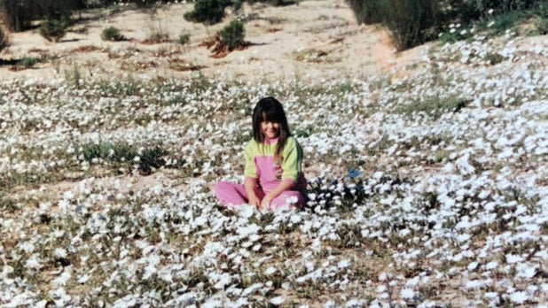 Shanna, as a child, sitting among white flowers in a natural setting with trees and rocks.